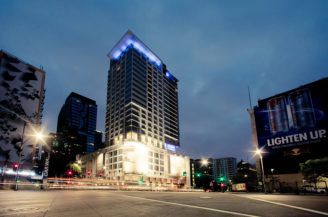 A tall, modern building in Downtown Los Angeles is illuminated at night with bright lights on its facade and a glowing blue top. Surrounding it are lower buildings with lit windows and two large billboards. The street in front, still quiet in the early morning, features light trails from passing vehicles.
