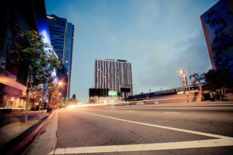 A wide city street at dusk in Downtown Los Angeles is flanked by tall buildings and a car wash on the right. The street is empty, and lights from passing cars create trails. The sky is cloudy, and the urban setting is captured with a long-exposure effect, giving a sense of motion.