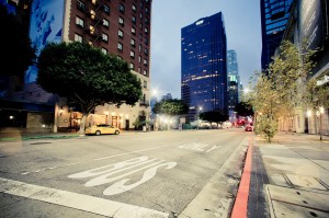 A quiet downtown Los Angeles street at dusk with tall buildings lining both sides. The road has "BUS ONLY" painted in white letters, and a lone yellow taxi is parked by the sidewalk. Trees with lush green leaves are seen along the street, under a partly cloudy sky with fading daylight.