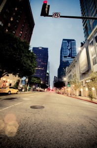Photo of a downtown Los Angeles street at dusk with tall buildings on both sides. Traffic lights are visible overhead, with one showing a red light. The street appears empty, and there are a few parked cars and trees lining the sidewalks. The sky has a faint blue hue.