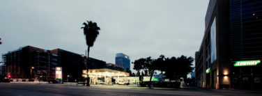 Wide-angle view of a bustling city street in Downtown Los Angeles at dusk, featuring a gas station on the left with a Shell logo, adjacent to a Subway restaurant on the right. A tall palm tree stands beside the gas station. Buildings and a high-rise tower are visible in the background.
