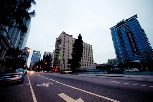 A wide street in Downtown Los Angeles with a bike lane, tall buildings on either side, and a few cars parked along the curb. The sky is overcast, and trees are visible alongside the buildings. One of the buildings has a large vertical "STAYINGLEVE.COM" banner, capturing an early morning vibe.