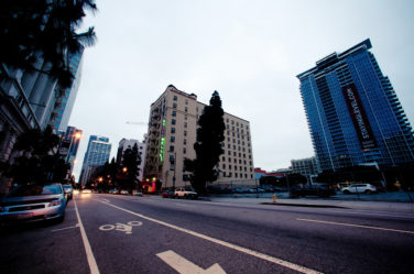 A wide street in Downtown Los Angeles with a bike lane, tall buildings on either side, and a few cars parked along the curb. The sky is overcast, and trees are visible alongside the buildings. One of the buildings has a large vertical "STAYINGLEVE.COM" banner, capturing an early morning vibe.