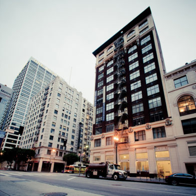 Early morning street-level view of downtown Los Angeles features tall buildings, including a prominent brown building with fire escape ladders on its facade. A truck is parked in front, and surrounding structures display various architectural styles. The sky is overcast.
