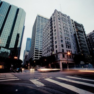 A city street in Downtown Los Angeles during the early morning or evening with tall office buildings and hotels lining the sidewalks. The crosswalk and streetlights are visible, and light trails from passing vehicles indicate motion in the otherwise quiet scene.