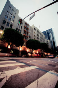 An early morning view of a downtown Los Angeles street features a multi-story building with large rectangular windows and an art-deco style facade. Trees line the sidewalk, lit by streetlights, while a street sign for Olive Way is visible above. Blurred motion of vehicles passing by is seen in the foreground.