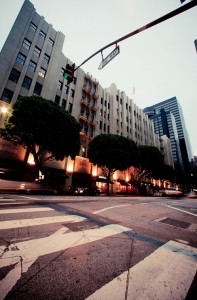 An early morning view of a downtown Los Angeles street features a multi-story building with large rectangular windows and an art-deco style facade. Trees line the sidewalk, lit by streetlights, while a street sign for Olive Way is visible above. Blurred motion of vehicles passing by is seen in the foreground.