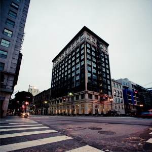 A tall, black high-rise building with large windows stands prominently at a street corner in Downtown Los Angeles. The intersection is marked by a zebra crossing and city lights, with cars passing by. Surrounding the building are other structures, creating an urban cityscape in the early morning.