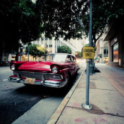A vintage red Ford car, an iconic vieille américaine, is parked on an urban street next to a "Loading Only" sign. The street is lined with tall buildings and trees, with shadows cast on the car and sidewalk. The area appears quiet and deserted.