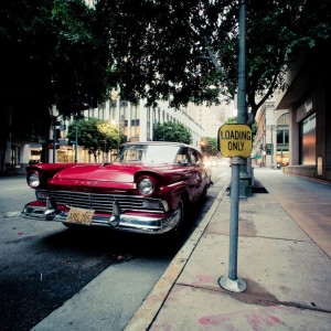 A vintage red Ford car, an iconic vieille américaine, is parked on an urban street next to a "Loading Only" sign. The street is lined with tall buildings and trees, with shadows cast on the car and sidewalk. The area appears quiet and deserted.