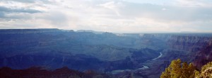 A panoramic view of the Grand Canyon under a partly cloudy sky. The vast landscape showcases deep, winding canyons, rugged cliffs, and a river flowing through the bottom. Captured with an XPAN camera, the scenery is bathed in soft, natural light, highlighting the canyon's layers and textures.