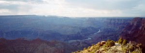 Wide panoramic view of the Grand Canyon showcasing its vast rocky landscape with layers of cliffs and a winding river below, captured beautifully in XPAN format. The sky is partly cloudy, casting shadows and light across the canyon, while vegetation is visible in the foreground.