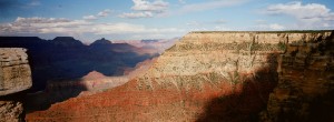 Panoramic view of the Grand Canyon, captured with an XPAN, showcasing its layered rock formations. The canyon walls display various shades of red, orange, and brown under a partly cloudy sky. Shadows accentuate the depth and rugged terrain of the canyon, highlighting its vast expanse.