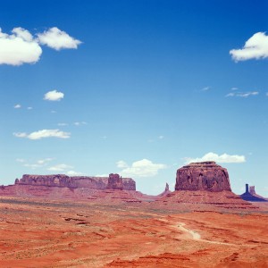 A vast desert landscape, reminiscent of Monument Valley, features towering red rock formations under a clear blue sky. Scattered white clouds dot the horizon. The predominantly red terrain, sparse in vegetation, is captured through the eye of the Hasselblad, with a winding dirt road cutting through this remote, untouched area.