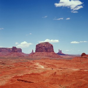 A vast desert landscape features towering red rock formations under a clear blue sky with a few scattered clouds. The rugged terrain with sparse vegetation stretches into the distance, revealing more rock structures reminiscent of Monument Valley, ideal for capturing stunning photography with a Hasselblad camera.