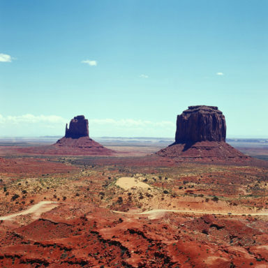 A vast desert landscape resembling Monument Valley features two large, distinct rock formations rising from the red sandstone terrain under a clear blue sky. Sparse shrubbery dots the arid ground, and a dirt road winds through the scene—perfect for some breathtaking Hasselblad photography.