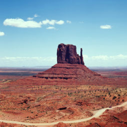 A towering butte stands prominently in the reddish desert landscape of Monument Valley, captured through the precise lens of a Hasselblad camera, under a clear blue sky with a few scattered white clouds. Sparse vegetation dots the surrounding area, offering an eye-pleasing contrast.
