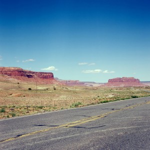A wide view of a desert landscape in Monument Valley with red rock formations under a clear blue sky. The foreground features an asphalt road with visible cracks and faded yellow lines. Sparse vegetation is scattered across the sandy and rocky terrain, perfect for Hasselblad photography enthusiasts.