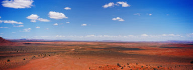A vast desert landscape stretches under a bright blue sky dotted with scattered white clouds. The arid terrain of Monument Valley is a mix of red and brown hues, with sparse vegetation. The horizon appears to merge with the distant mountains, forming a picturesque backdrop that catches the eye.