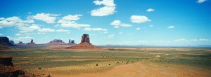 A panoramic view of a vast desert landscape in Monument Valley with large, isolated red rock formations under a bright blue sky dotted with scattered white clouds. The terrain is mainly flat with sparse vegetation, illustrating the expansive and arid nature of the scene captured by XFAN.