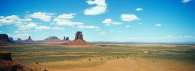A panoramic view of a vast desert landscape in Monument Valley with large, isolated red rock formations under a bright blue sky dotted with scattered white clouds. The terrain is mainly flat with sparse vegetation, illustrating the expansive and arid nature of the scene captured by XFAN.