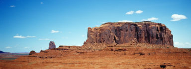 A vast desert landscape reminiscent of Monument Valley features a large, rugged rock formation under a clear blue sky. The formation has steep cliffs and a flat top, with a distant butte visible to the left. Sparse clouds dot the sky, adding depth to this arid yet mesmerizing XPAN-worthy scene.