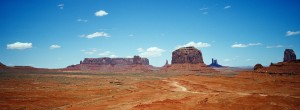 Panoramic view of Monument Valley showcasing vast, arid desert terrain with distinctive, towering sandstone buttes under a clear blue sky scattered with a few small clouds. A winding dirt road cuts through the red landscape, capturing the eye and inviting exploration with an XPAN-like breadth.