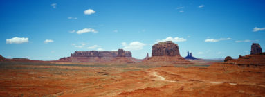Panoramic view of Monument Valley showcasing vast, arid desert terrain with distinctive, towering sandstone buttes under a clear blue sky scattered with a few small clouds. A winding dirt road cuts through the red landscape, capturing the eye and inviting exploration with an XPAN-like breadth.