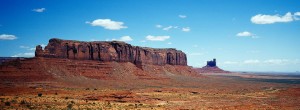An expansive desert landscape reminiscent of Monument Valley features towering red rock formations under a bright blue sky with scattered clouds. A large plateau dominates the left side, with a tall, isolated rock formation in the distance to the right. Sparse vegetation covers the ground, creating an XPAN-like panorama.