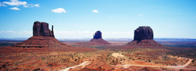 A vast desert landscape with three prominent rock formations, known as buttes, rising from the ground in Monument Valley. The sky is clear and blue, with a few scattered clouds. The scene is expansive, with the red and orange hues of the desert stretching to the horizon—a perfect setting for XPAN photography.