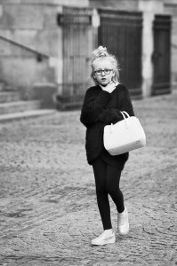 A young girl with glasses, wearing a dark sweater, leggings, and light shoes, walks on a cobblestone street holding a white handbag. Elle a tout d'une grande in this black and white image.