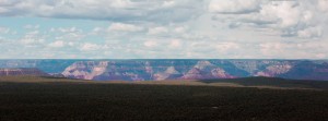 A panoramic view of the Grand Canyon, captured with an XPAN camera, showcasing a vast landscape with deep gorges and rugged cliffs under a partly cloudy sky. The canyon's colorful layers of rock formations are visible in the distance, while a forested area in the foreground contrasts with the arid scenery.