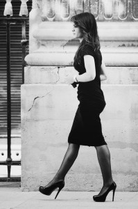 A woman in mode, dressed in a fitted black dress and hauts talons, walks confidently on a city sidewalk beside a large stone building, seen in profile against an ornate urban backdrop. The image is in black and white.