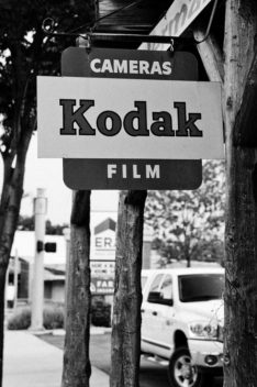 Black and white image of a vintage Kodak sign that reads "CAMERAS Film" on a wooden post. The sign, photographed by Terry Alderman, is juxtaposed against a blurred background of trees, the streets of Kanab, and a white pickup truck.