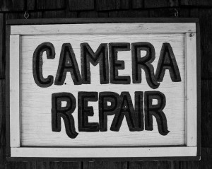 A black and white image of a weathered wooden sign that reads "CAMERA REPAIR" in bold, block letters. The sign, framed with a simple wooden border, hangs against a dark background. This vintage piece evokes the charm of Terry Alderman's photo boutique in Kanab, Utah.