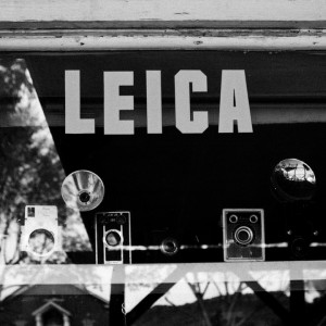 Black and white photo of a boutique photo store window display in Kanab, Utah, featuring vintage Leica cameras and accessories. The word "LEICA" is prominently displayed at the top of the window. Reflections of trees and houses are visible in the glass, adding to its timeless charm.