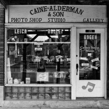 Black and white photo of a storefront with signage reading "Caine-Alderman & Son Photo Shop Studio Gallery." The display window showcases cameras and photography equipment. The door has signs for Leica, Rollei, Bogen, music notes, and "Center Street Music." Owned by Terry Alderman, this boutique photo shop adds a charming touch to Kanab.