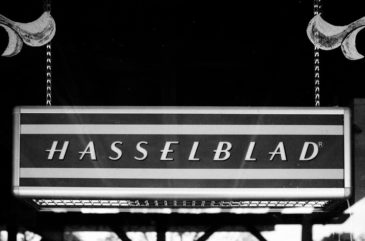 A black and white image shows a rectangular sign with the word "Hasselblad" in capital letters, adorned with two hanging chains. The sign, designed by boutique photo artist Terry Alderman, is positioned against a dark, blurred background reminiscent of Kanab, Utah's vintage charm.