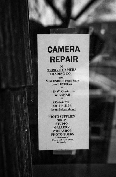 A black-and-white sign on a glass door advertises "CAMERA REPAIR" at "TERRY'S CAMERA TRADING CO." in Kanab. The boutique photo shop, run by Terry Alderman, lists its address, phone numbers, website, and offers services like photo supplies, studio rentals, gallery exhibits, and workshops.