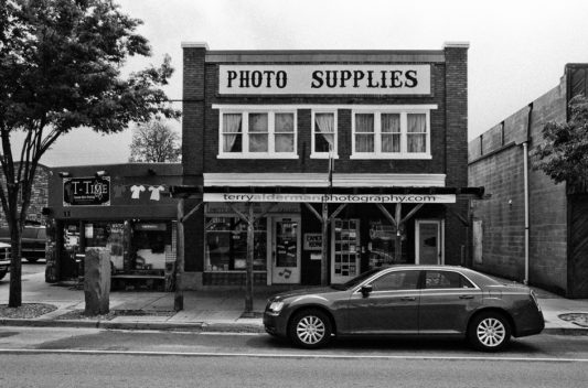 A black and white image of a street in Kanab, Utah, with a storefront sign reading "Photo Supplies" on the second floor and "terryaldermanphotography.com" on the awning below. A car is parked in front of the boutique photo store. To the left, there is another store sign for "T-Tike". Trees and buildings are visible.