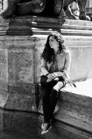 Carole Desinaud A young woman with long hair sits alone on a stone ledge by a large monument, looking to the side. She wears a jacket, dark pants, and sneakers. The black and white photo evokes the timeless style of Carole Desinaud.