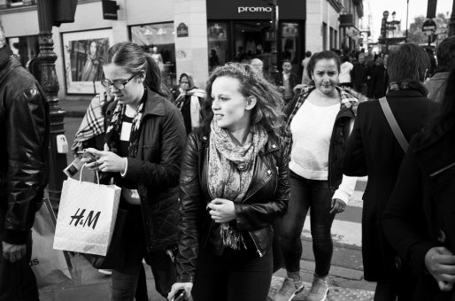 Foule de fin de semaine A group of people walk along a busy city street. Amid the fin de semaine foule, two women in front wear jackets and scarves; one carries an H&M shopping bag while the other looks ahead. Storefronts and more people fill the background.