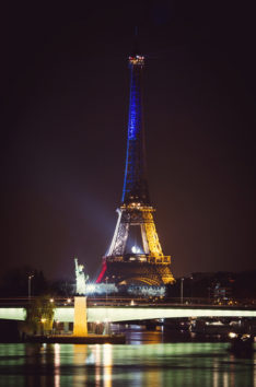 Nighttime view of the tour Eiffel illuminated in bleu blanc rouge lights. In front, a smaller replica of the Statue of Liberty stands on a small island surrounded by water, with a bridge and reflections on the water in the foreground.
