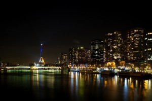 Nighttime cityscape of Paris featuring the tour Eiffel illuminated in blue. The Seine River reflects the city's lights, with high-rise buildings lining the riverbank. The scene captures a mix of modern architecture and historic landmarks, all under the hues of bleu blanc rouge.