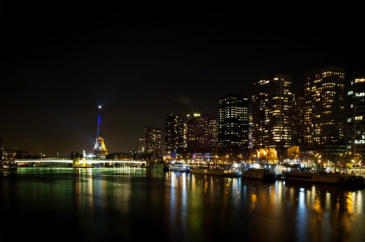 Nighttime cityscape of Paris featuring the tour Eiffel illuminated in blue. The Seine River reflects the city's lights, with high-rise buildings lining the riverbank. The scene captures a mix of modern architecture and historic landmarks, all under the hues of bleu blanc rouge.