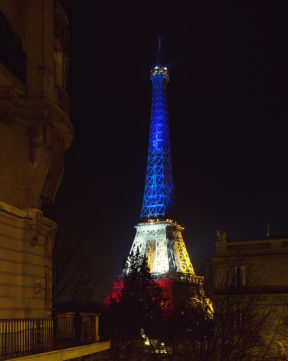 The Eiffel Tower illuminated at night with the colors blue, yellow, and red against a dark sky. The colors create a gradient effect from top to bottom. Surrounding buildings and trees are dimly visible in the foreground, while white lights subtly highlight architectural details.