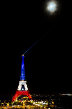 The Eiffel Tower is illuminated in bleu, blanc, rouge lights against a dark night sky, symbolizing the colors of the French flag. A bright moon is visible above, partially obscured by some clouds, casting a soft glow over this iconic Eiffel scene.