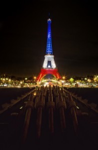 Night view of the Eiffel Tower illuminated with the colors of the French flag, Bleu Blanc Rouge. Bright lights surround the Eiffel, reflecting on the dark waters of the fountain in the foreground, where cannons can be seen pointing toward the tower.