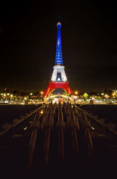 Night view of the Eiffel Tower illuminated with the colors of the French flag, Bleu Blanc Rouge. Bright lights surround the Eiffel, reflecting on the dark waters of the fountain in the foreground, where cannons can be seen pointing toward the tower.