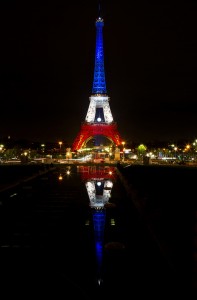 Night view of the tour Eiffel illuminated with bleu blanc rouge lights, reflecting the French flag colors. The tower's reflection is visible in a calm water surface below. Surrounding city lights and dark sky frame the iconic structure.